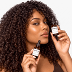 A woman with voluminous dark brown curly hair holds two amber glass dropper bottles labeled 'BATANA OIL' against a clean white background.