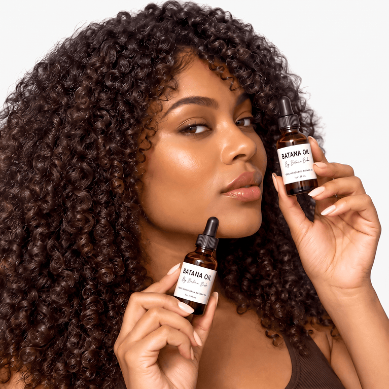 A woman with voluminous dark brown curly hair holds two amber glass dropper bottles labeled 'BATANA OIL' against a clean white background.